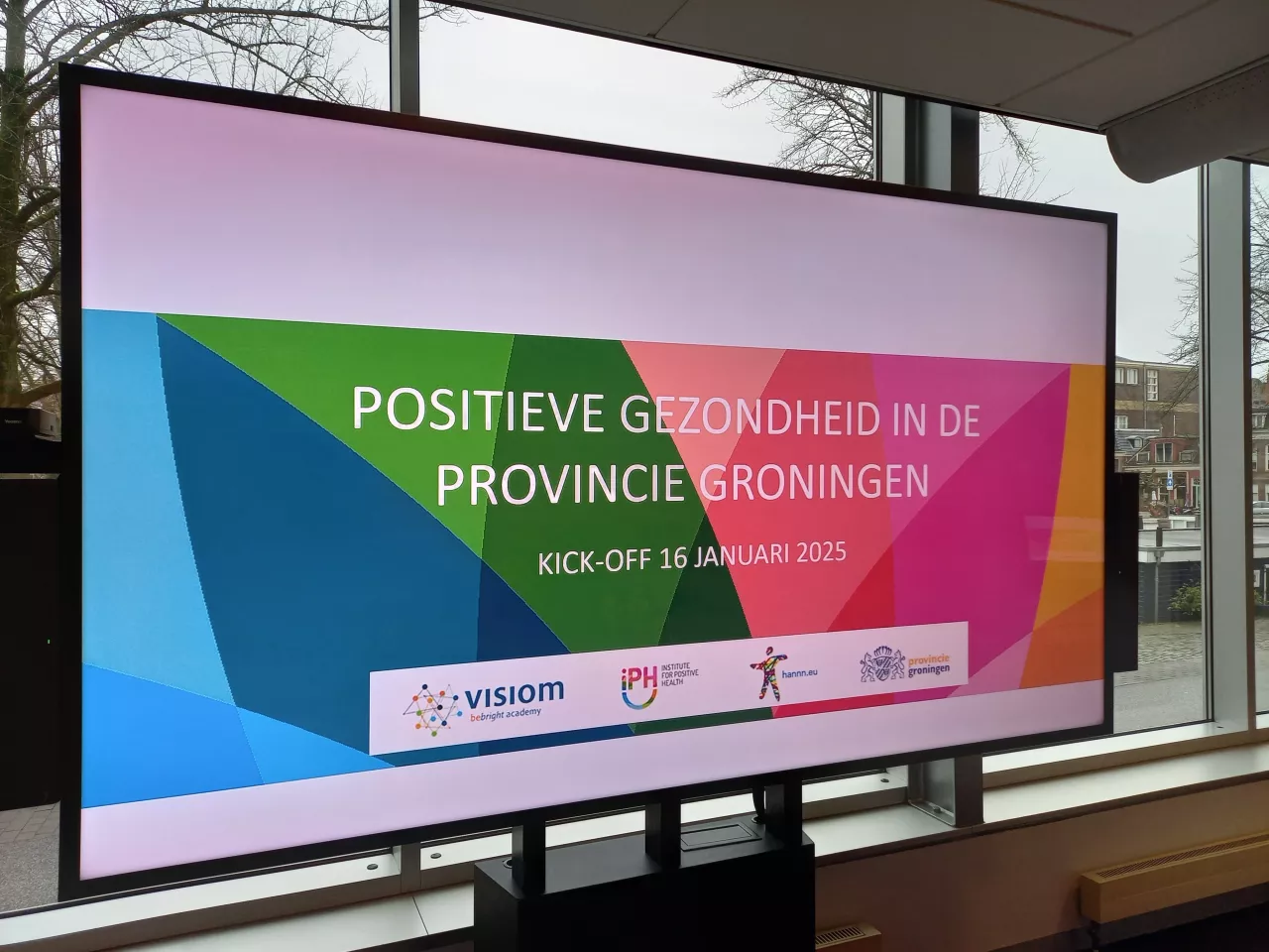 Tv scherm de de titel van de bijeenkomst erop: 'Positieve Gezondheid in de Provincie Groningen' en de logo's van de betrokken partners.