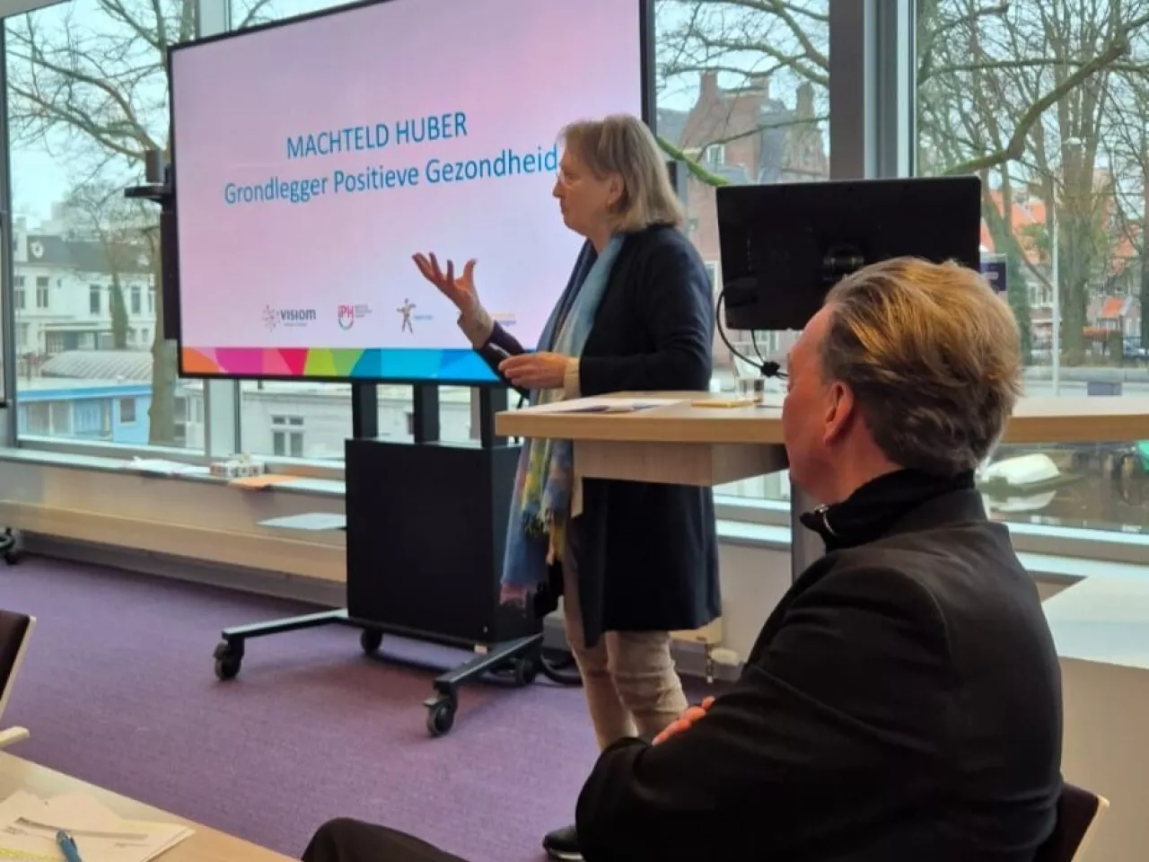 Machteld Huber van Positieve Gezondheid aan het spreken naar de groep voor het tv scherm, met gedeputeerde Tjeerd van Dekken in het beeld.